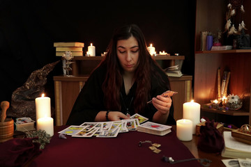 A witch is fortune teller in black robe wonders using tarot cards on table. Magical ritual. White candles, aroma stick, ancient books, runes, amulets on table. Occult, esoteric and divination concept.