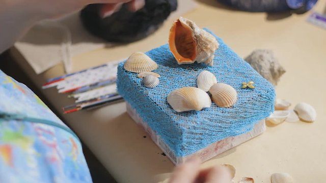 Female artist is making a design on the top cover of souvenir blue paper box with various sea shells
