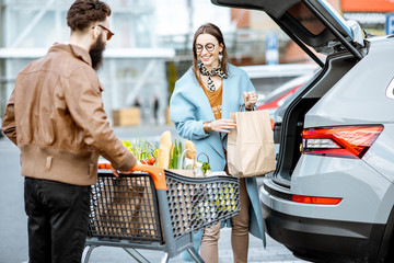 Young stylish couple with shopping cart full of fresh food, packing products into the car on the...