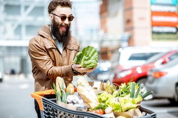 Portrait of a stylish man with shopping cart full of fresh and healthy food on the outdoor parking near the supermarket