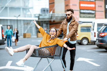 Young stylish coupe having fun riding with shopping cart on the outdoor parking near the supermarket