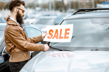 Stylish bearded salesman with sales plate near the cars on the open ground of a dealership