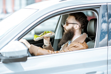Naklejka premium Stylish bearded man having a snack with tasty sandwich while driving a car in the city