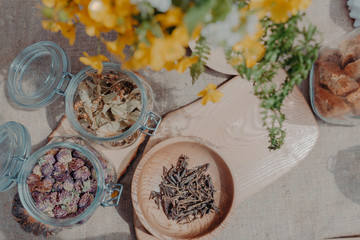 Tea herbs (red clever, sagan daylya, linden flowers) in bowl and jar on table with yellow flowers and jars with cookies near. Grey background, copy space. Horizontal orientation. Top view.