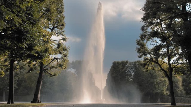 Hannover, Germany. A high jet of water from the fountain spouting from a bowl set on the ground in the setting sun. Against the background of green trees in the park. The concept of saving natural
