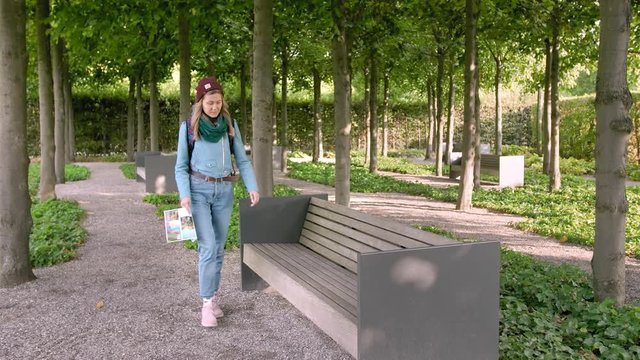 Hannover, Germany. A young female tourist with a map in her hands and a backpack sits on a stone bench in the park. Opens a paper map and looks the way in it. In the frame a lot of trees. Slow Motion.