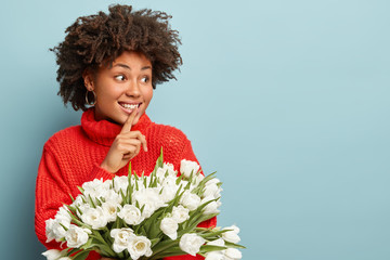 Photo of curly beautiful woman looks joyfully aside, makes silence gesture, dressed in red winter sweater, tells secret who gave white tulips, gossips with friend, isolated on blue. Lady holds flowers