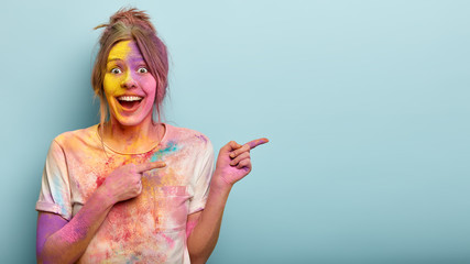 Horizontal studio shot of happy European woman points at side, covered with Holi colors, wears...