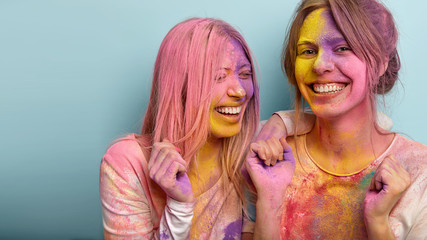 Obraz premium Cropped image of joyful young women laugh and clench fists, have fun together on Holi festival, throw colorful paints at each other, express positive emotions, pose in studio over blue background
