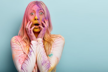 Intense shocked blonde European woman smeared with multicolored powder, gasps and opens mouth, holds hands near face, celebrates Holi run, impressed to see many participants. Copy space aside