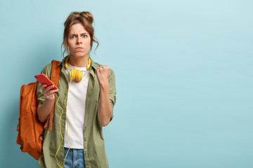 Image of angry millennial woman clenches fists and looks with irritation at camera, annoyed by noisy people who disturb her, wants to listen music in peaceful atmosphere, stands with bag indoor.