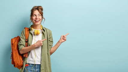 Positive European female teenager uses modern technologies for entertainment, points aside on free space, dressed in loose shirt and jeans, carries backpack, promotos something, poses indoor - obrazy, fototapety, plakaty