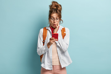 Image of astonished young female addicted to internet, networks via cell phone, surprised to have limits, has dark combed hair, focused at screen, carries backpack, isolated on blue background.