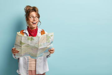Cheerful European woman has interesting trip, looks aside, holds map, checks route or location,...