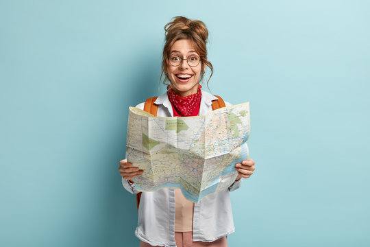 Happy female traveler holds map, checks route for destination, has trip across Europe during holidays, carries backpack, wears round spectacles, isolated on blue wall, plans tour, checks location