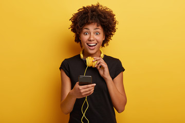 Happy enthusiastic female holds smart phone device connected to stereo headphones, smiles...