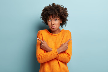 Pick one thing only. Clueless frustrated dark skinned female model crosses hands over chest, points at different empty corners, has hesitant facial expression, wears orange jumper, stands indoor