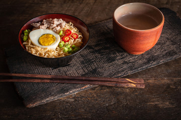 Instant noodle with pork, egg and vegetables on black bowl on the wood table there are chopstick and drinking glass placed around.