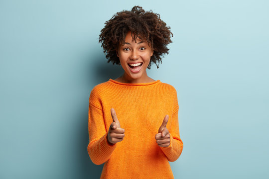 Joyous pretty African American lady makes finger gun gesture at camera, expresses choice, smiles broadly, dressed in orange jumper, isolated over blue background, says you are chosen. Bang, bang