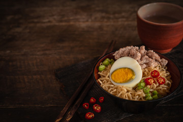 Instant noodle with pork, egg and vegetables on black bowl on the wood table there are chilli, chopstick and drinking glass placed around.