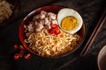 Instant noodle with pork, egg and vegetables on black bowl on the wood table there are chilli, chopstick, instant noodle in black plate and drinking glass placed around.