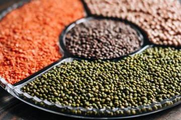 Various legumes red lentils, mung beans and chickpeas in a glass plate on a wooden table close up