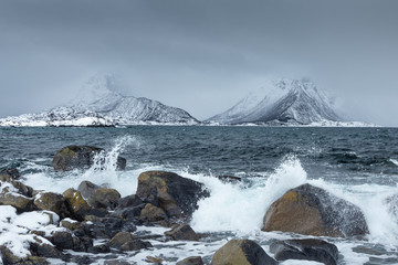Fototapeta na wymiar Waves crushing ont he rocks on a beach from Lofoten Islands, Norway. Spectacular snowy mountains in the background. Moody witner day