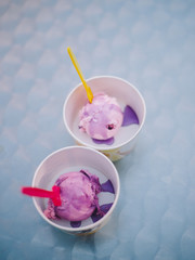 Close-up of two cups of lilac ice cream with syrup and color spoons stand on the table on a blue blurred background. Selective focus
