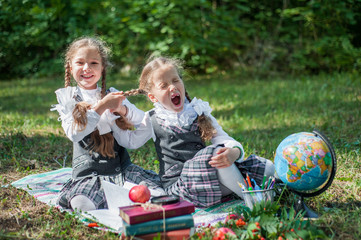Two girls in school uniform sit on the grass with books, pencils, a globe and an apple