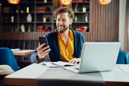 Young Caucasian Businessman Dressed Smart Casual Using Smart Phone While Sitting In Cafe. On The Desk Laptop.