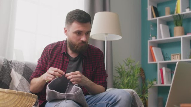 Medium shot of man sitting on sofa and watching knitting tutorial on laptop