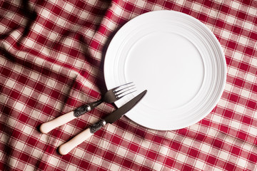 Empty white plate, fork and knife on a checkered red napkin, top view. Image with copy space. Kitchen table with a towel and a plate - top view with copy space. 