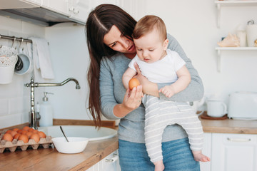 Young mother brunette woman with baby boy in arms cooking food in white modern kitchen at home