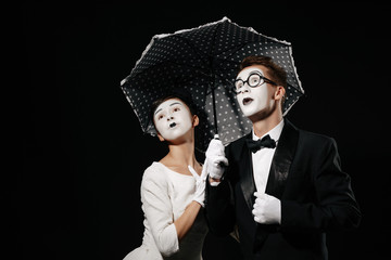 portrait of surprised couple mime with umbrella on black background. man in tuxedo and glasses and woman in white dress