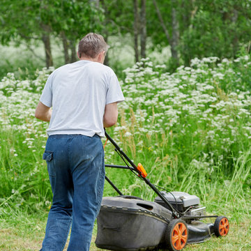 Gardener is working in the yard of a countryside cottage, he is using lawn mower.
