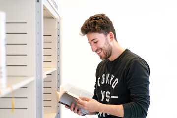 Young attractive green-eyed student smiling happily with a book in his hand in front of a bookshelf in the library