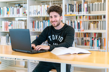 Young student sitting in front of the computer with books at the table happy smiling in the library