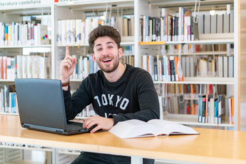 Young student sitting in front of the computer with books at the table happy with finger up high with success