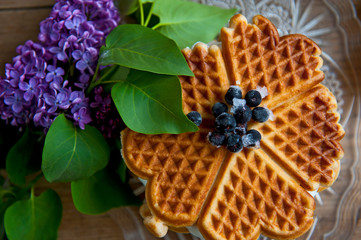 Homemade waffles with aronia, eggs and lilac flowers on the wooden table
