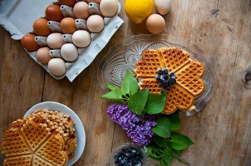 Homemade waffles with aronia, eggs and lilac flowers on the wooden table