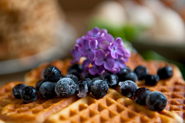 Homemade waffles with aronia, eggs and lilac flowers on the wooden table