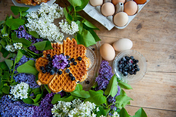 Homemade waffles with aronia, eggs and lilac flowers on the wooden table
