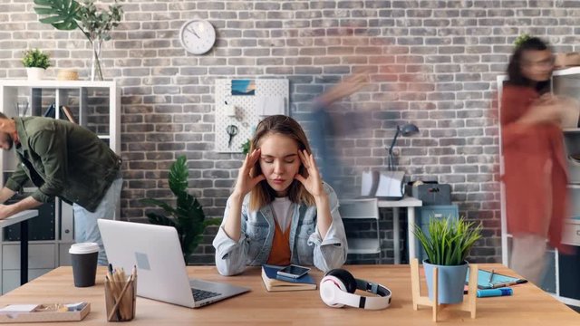 Zoom out time-lapse of tired young woman exhausted employee touching head in office feeling sick sitting at desk while busy coworkers are rushing around.