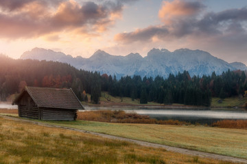 Wonderful misty morning with colorful sky over the mountain valley.  View over Geroldsee with wooden hut and Karwendel mountains at early morning in autumn, Bavaria, Germany.