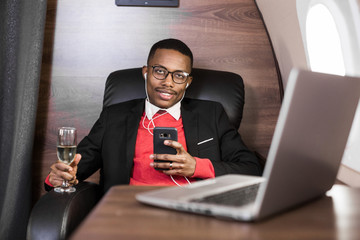 Fototapeta na wymiar Attractive and successful African American businessman with glasses working on a laptop while sitting in the chair of his private jet