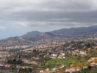 Fototapeta na wymiar aerial cityscape view of the outskirts of funchal in Madeira with farms and houses with mountains and cloudy sky in the distance