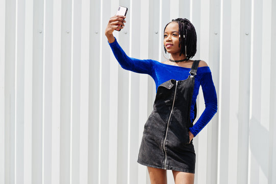 African woman in denim overalls skirt and blue dress posed against white steel wall and making selfie on phone. - Powered by Adobe