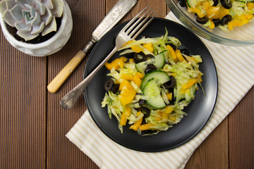 Fresh salad of black olives, iceberg cabbage, yellow bell pepper and cucumber, healthy food flat lay, natural light, wooden table.