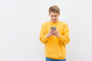 Fototapeta premium Close-up portrait of a guy in a yellow sweatshirt, standing against a white wall, enjoying a smartphone rejoicing. Happy young in casual clothing writes a message on the smartphone and smiles