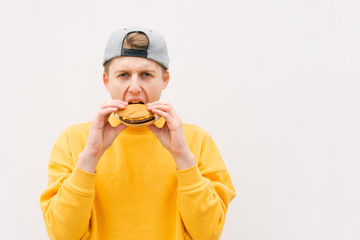 Hungry young man standing in the street on the background of a white wall and eating a sandwich. Student bites a sandwich and looks at the camera, isolated against the background of a white wall